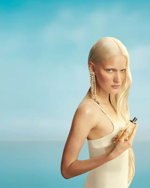 Nuevo banner de campaña de Mugler Starlicious les eaux gourmandes con una modelo con cabello rubio largo y ojos azules, con un vestido amarillo pálido, sosteniendo el frasco de la fragancia Pineapple Musk contra un fondo azul cielo suave. Las fragancias son deliciosas, coleccionables y combinables.